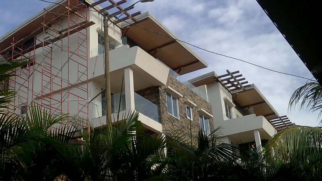 An apartment block mid-construction: white render, a stone-clad feature wall, glass balustrades and timber pergolas over the balconies, with scaffolding still in place.